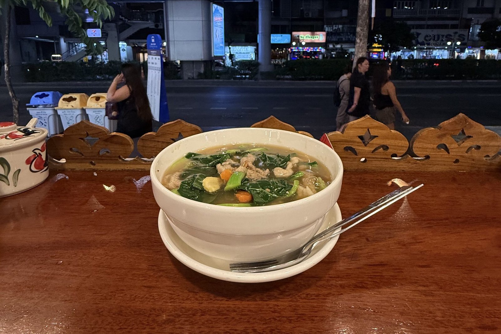A bowl of soup on a wooden table overlooking a Bangkok night street.